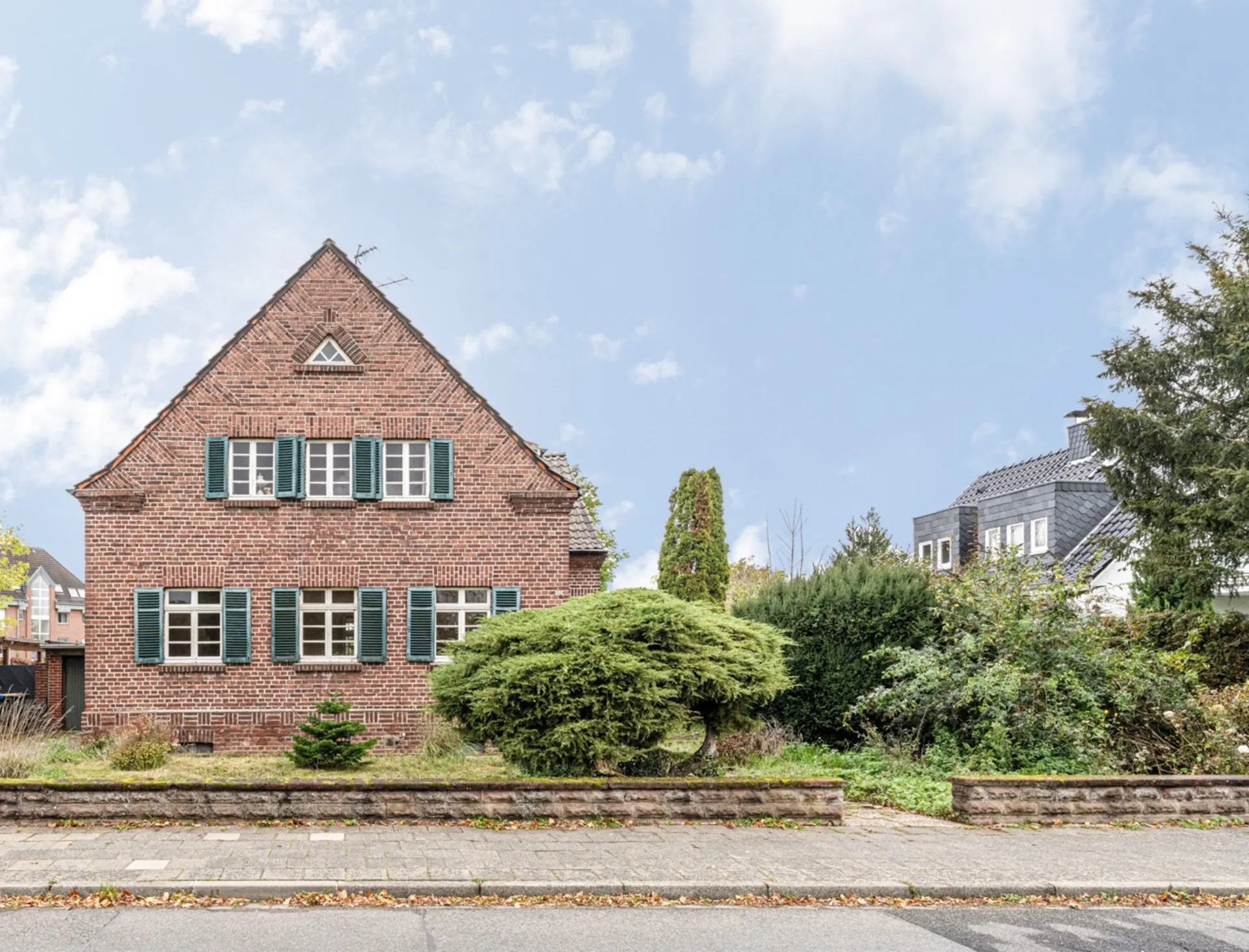 Backsteinhaus mit grünen Fensterläden, kleinem Vorgarten und Strauch vor dem Haus.