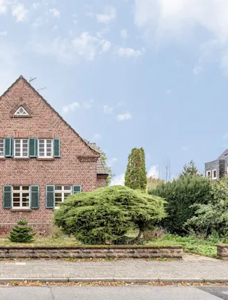 Backsteinhaus mit grünen Fensterläden, kleinem Vorgarten und Strauch vor dem Haus.