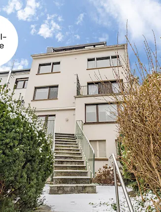 Mehrstöckiges Wohnhaus mit Treppe, umgeben von winterlichen Büschen, unter blauem Himmel.