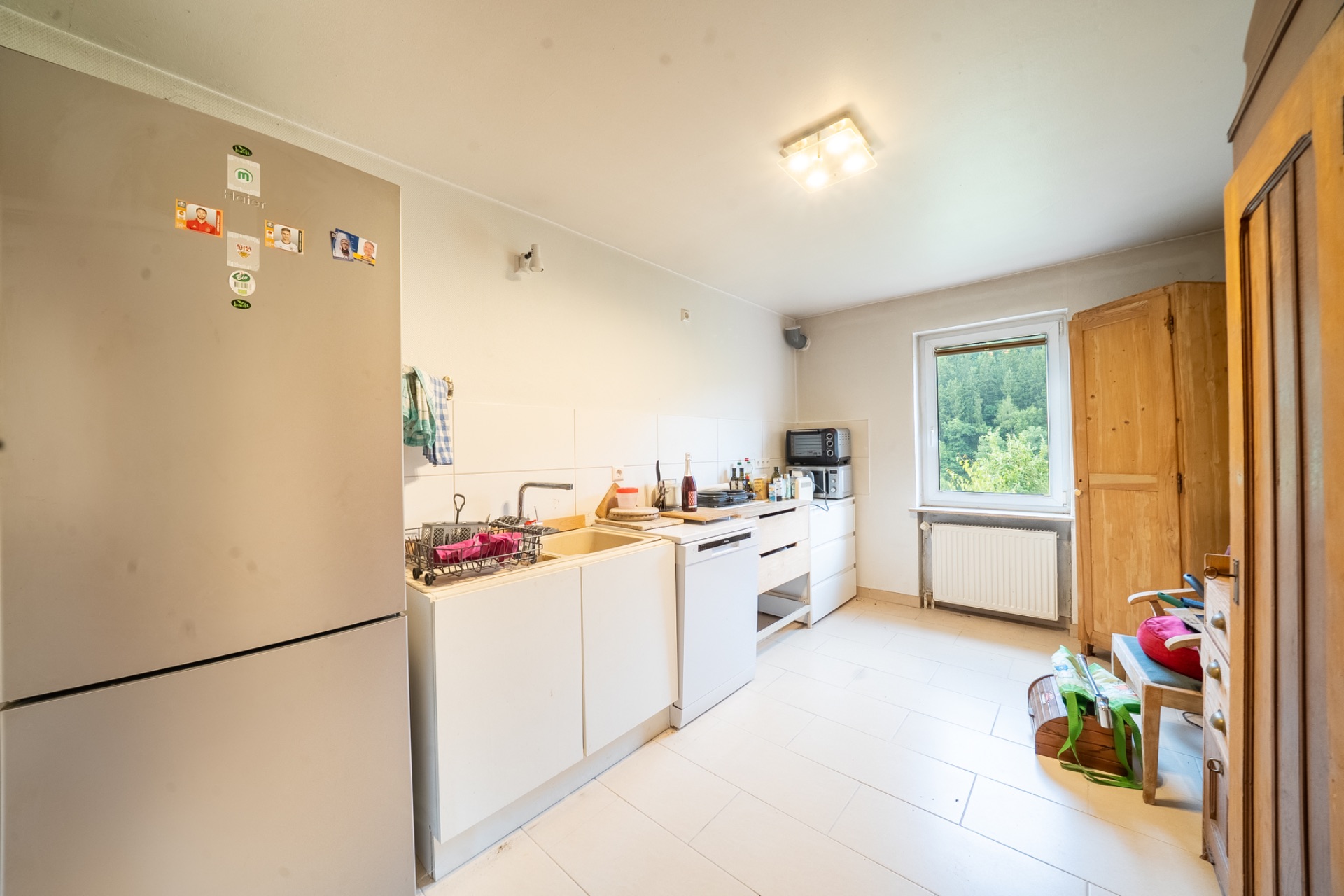 Helle Küche mit Kühlschrank, Herd, Spüle und Fenster mit Blick ins Grüne.