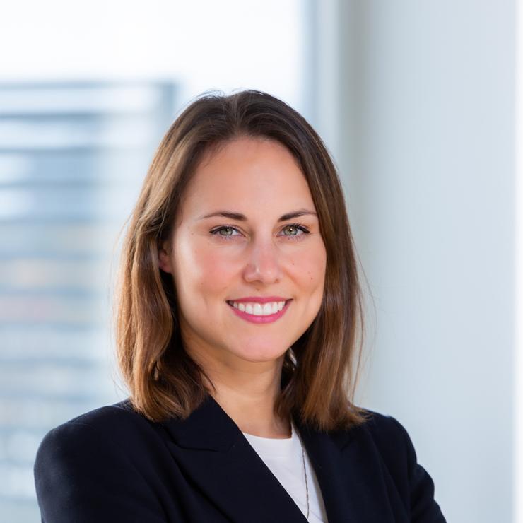 Frau mit braunen Haaren in dunklem Blazer steht lächelnd in modernem, hellen Raum.