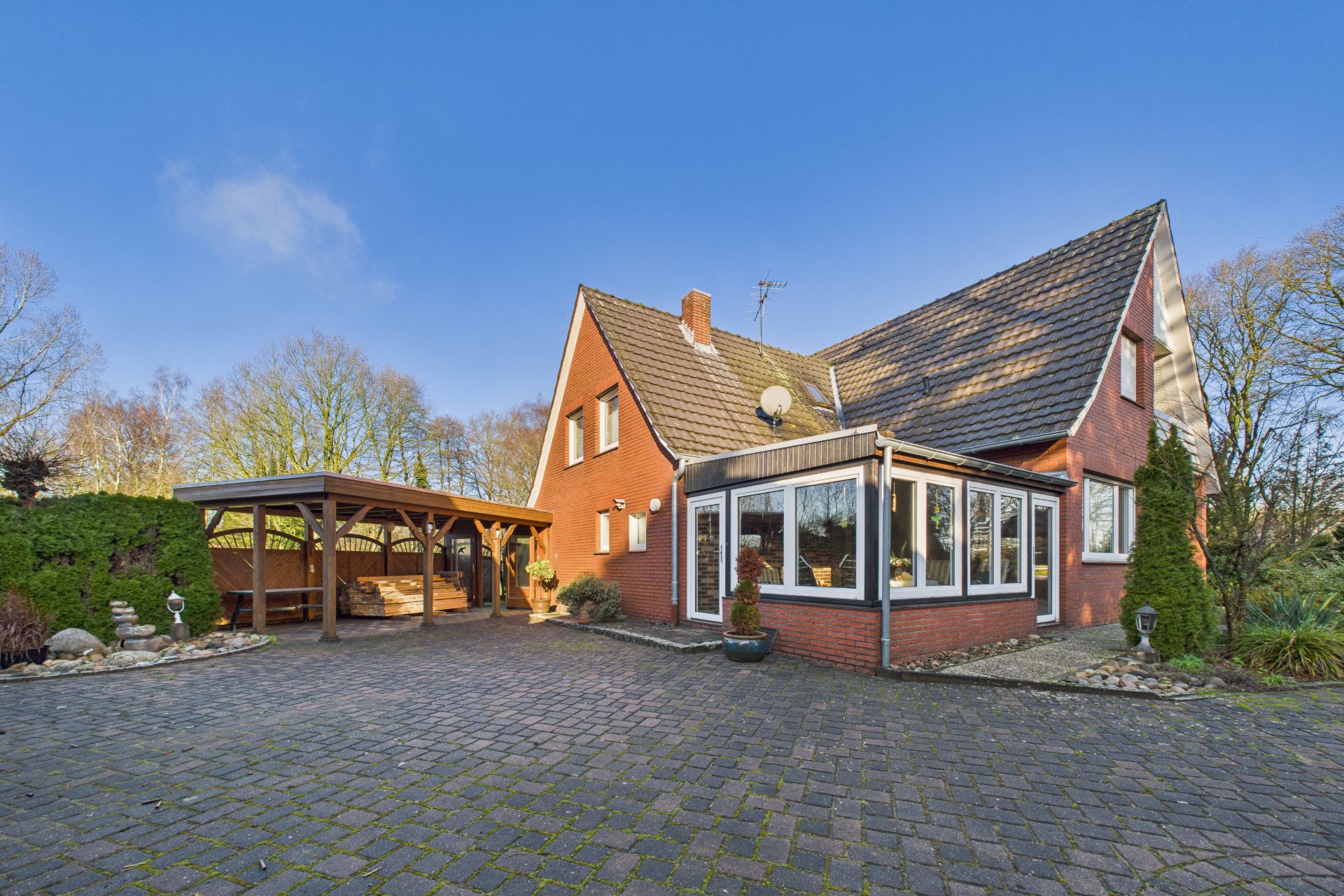 Backsteinhaus mit Wintergarten, Pflasterauffahrt und Carport, umgeben von Bäumen.