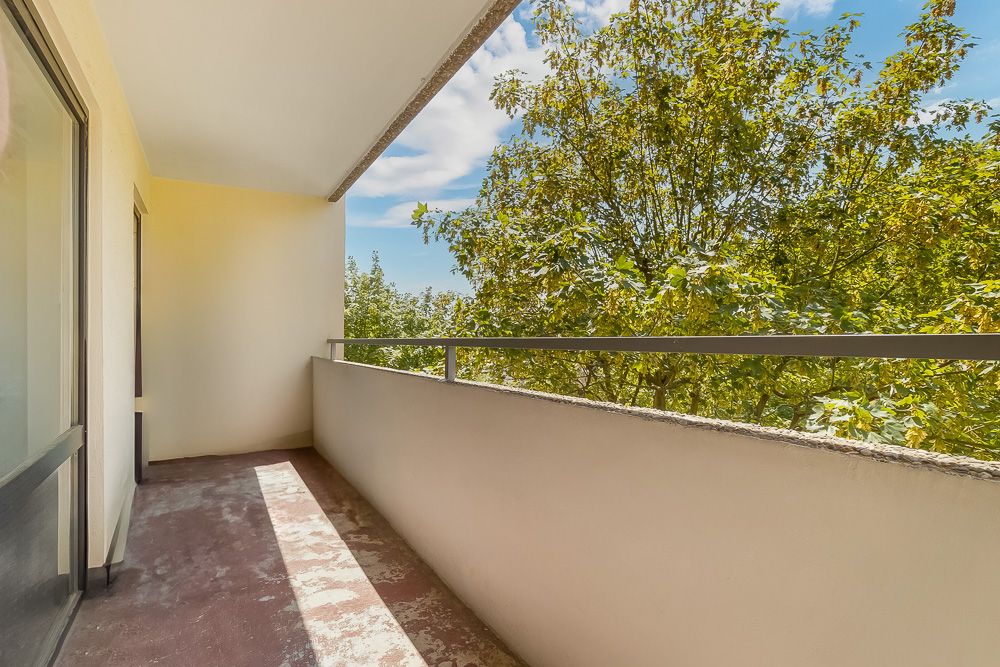 Überdachter Balkon mit bräunlichem Bodenbelag, Geländer und Blick auf grüne Bäume.