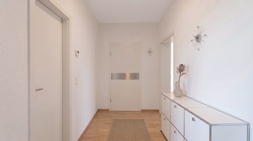 Heller Flur mit Holzboden, Kommode, Vase und geschlossener Tür am Ende.