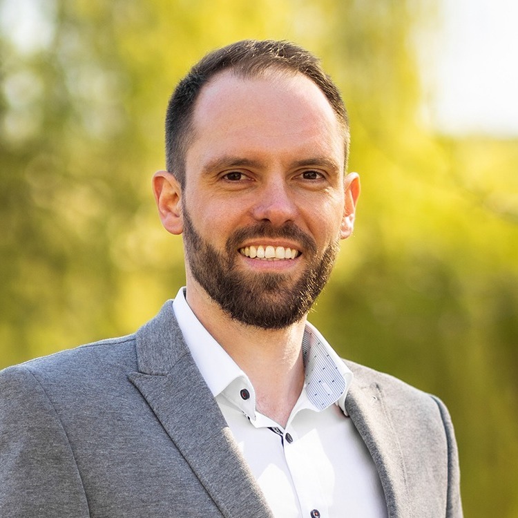 Ein Mann im grauen Sakko steht draußen vor unscharfem grünem Hintergrund und lächelt.