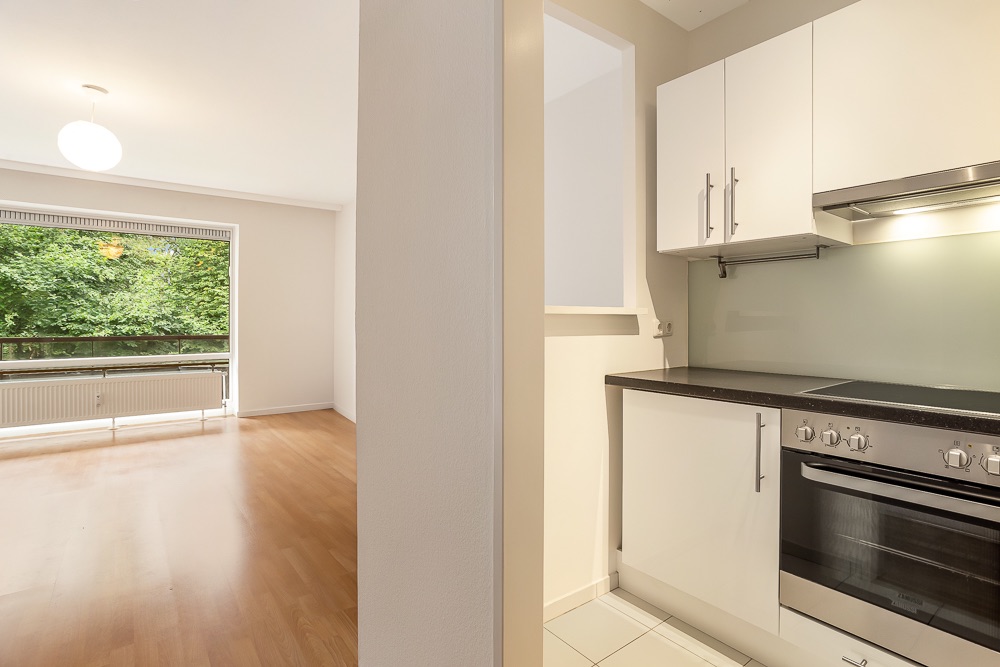 Helles Wohnzimmer mit Holzfußboden und große Fenster, daneben eine moderne Küche mit weißen Schränken.