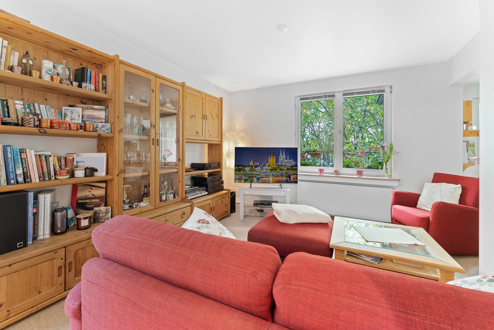 Wohnzimmer mit roten Sofas, Holzregalen, Fernseher und großem Fenster mit Blick ins Grüne.