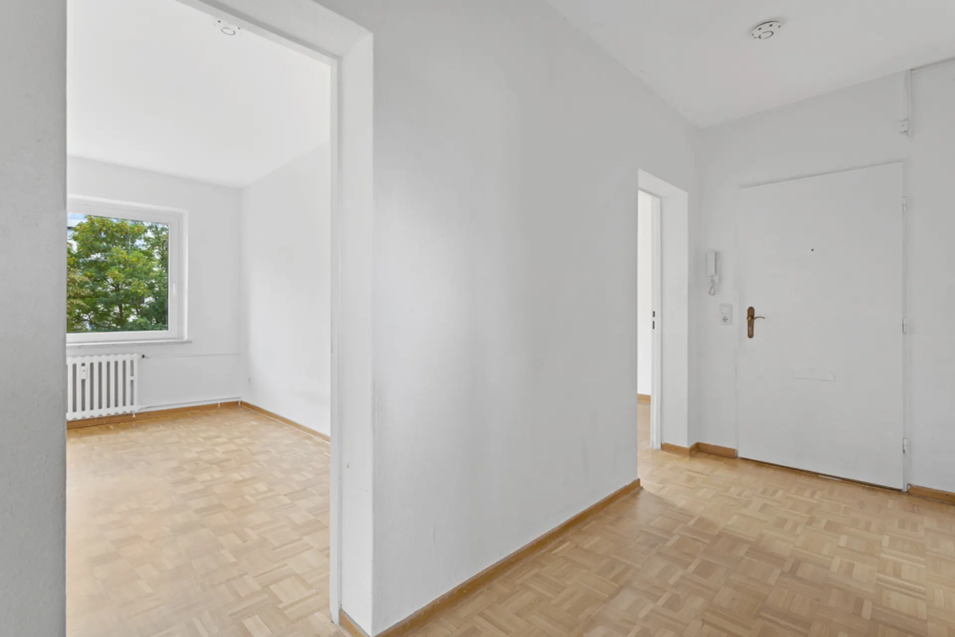 Heller Flur mit Parkettboden und Blick in ein Zimmer mit Fenster und Heizkörper.