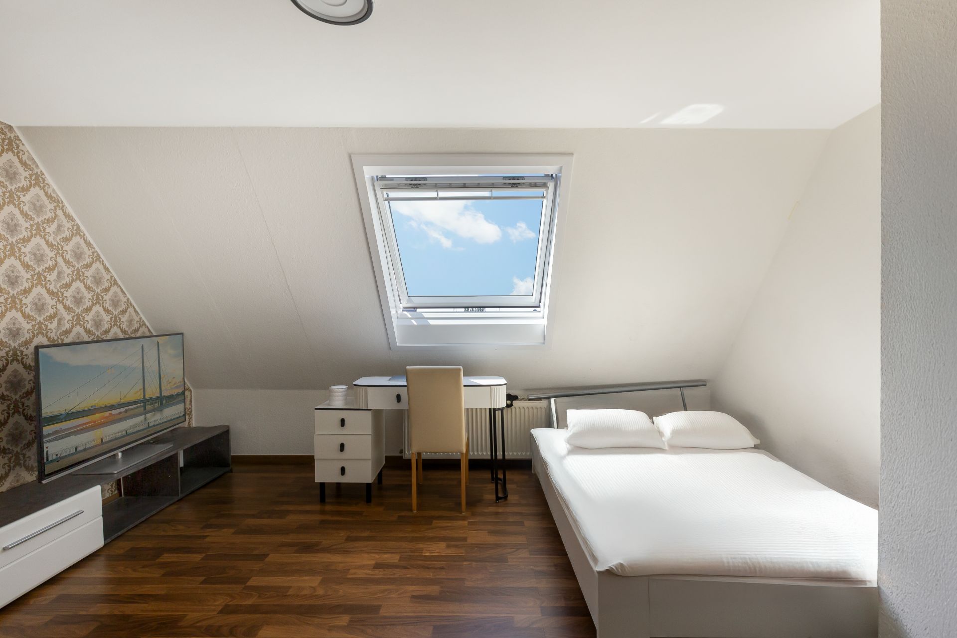 Helles Schlafzimmer mit Doppelbett, Schreibtisch, Fernseher und Dachfenster.