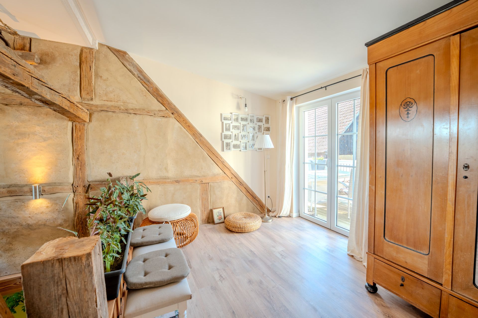 Helles Zimmer mit Holzbalken, großem Schrank, Sitzbank, Pflanzen und Glasflügeltür zum Balkon.