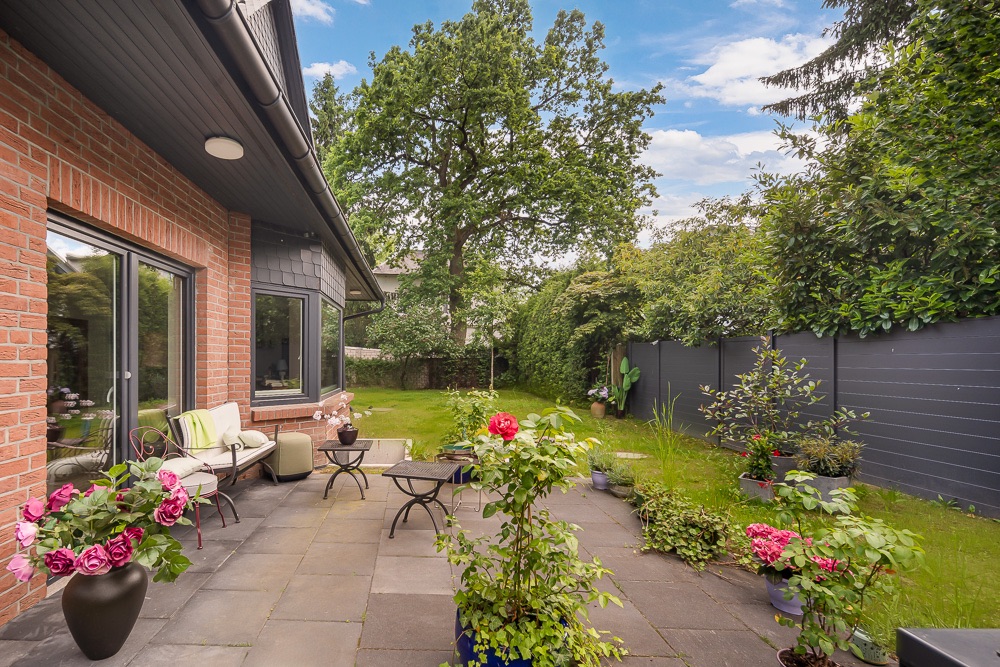 Terrasse mit Garten, Blumen in Töpfen, Sitzgelegenheiten und großem Baum im Hintergrund.