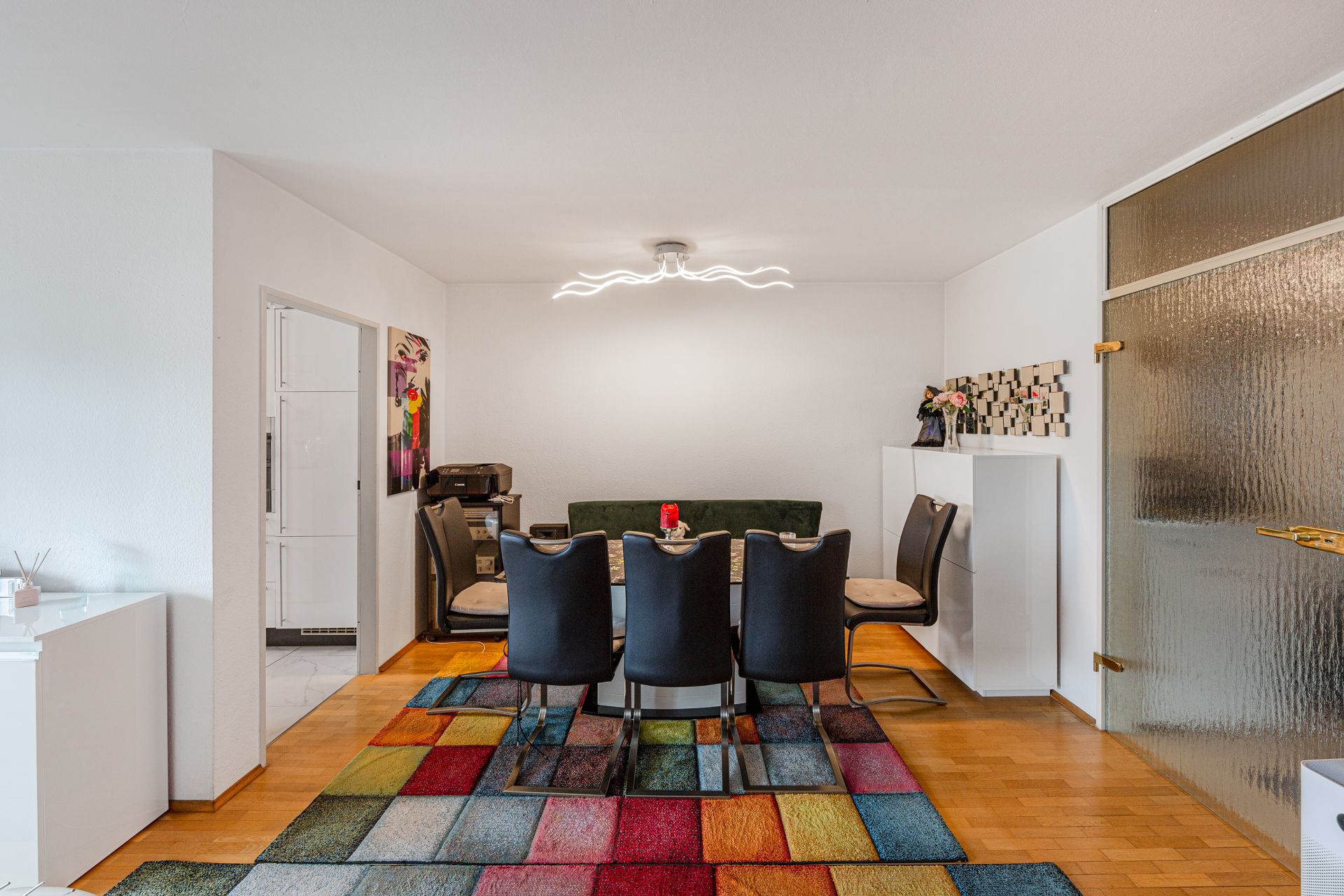 Esszimmer mit sechs schwarzen Stühlen, buntem Teppich und modernem Licht an der Decke.