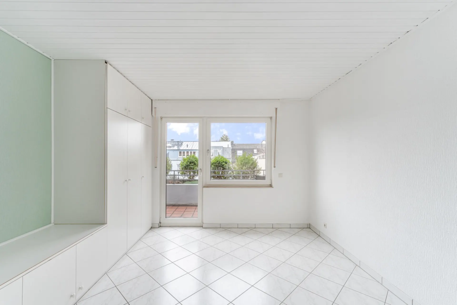 Heller Raum mit weißen Einbauschränken, Fliesenboden und Zugang zu einem Balkon.