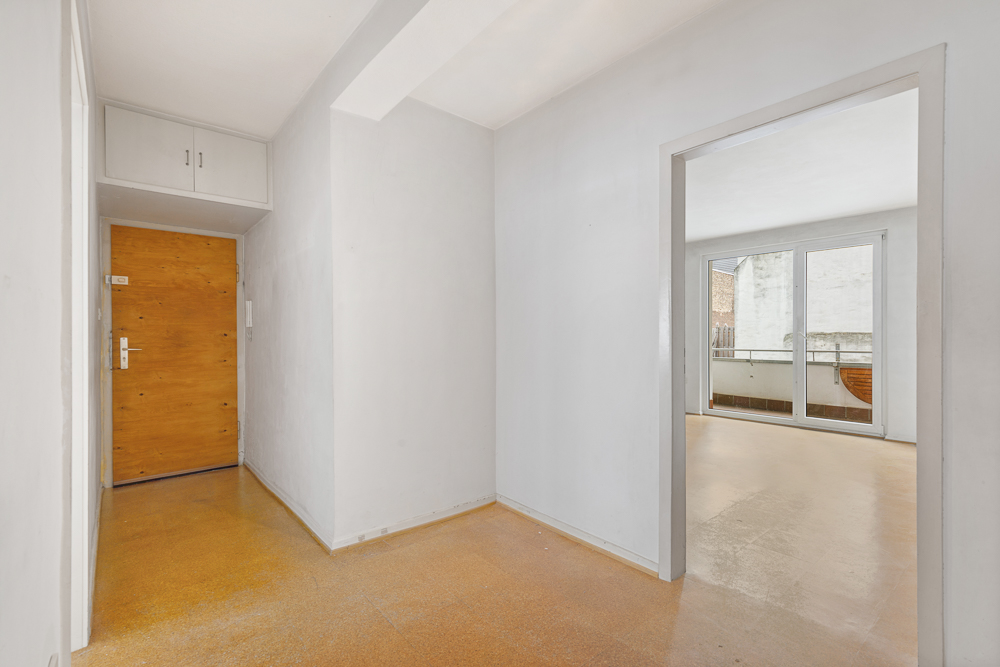 Heller Flur mit Holztür, weißer Wand und Zugang zu einem Zimmer mit großen Fenstern.