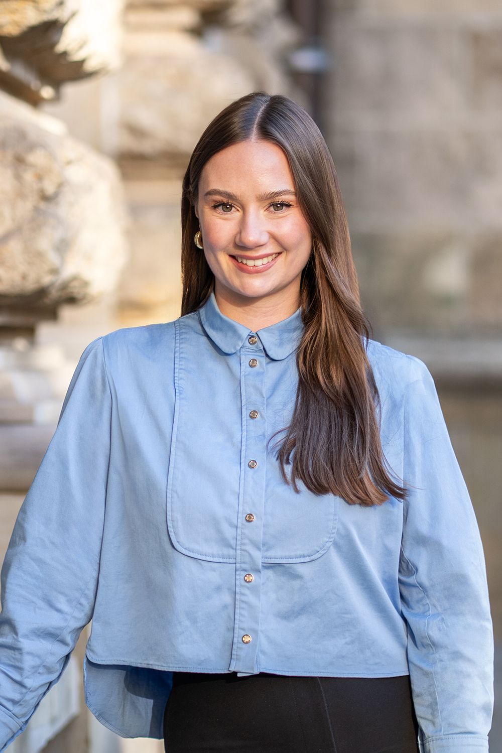 Eine Frau mit langem braunem Haar trägt eine hellblaue Bluse und steht draußen.