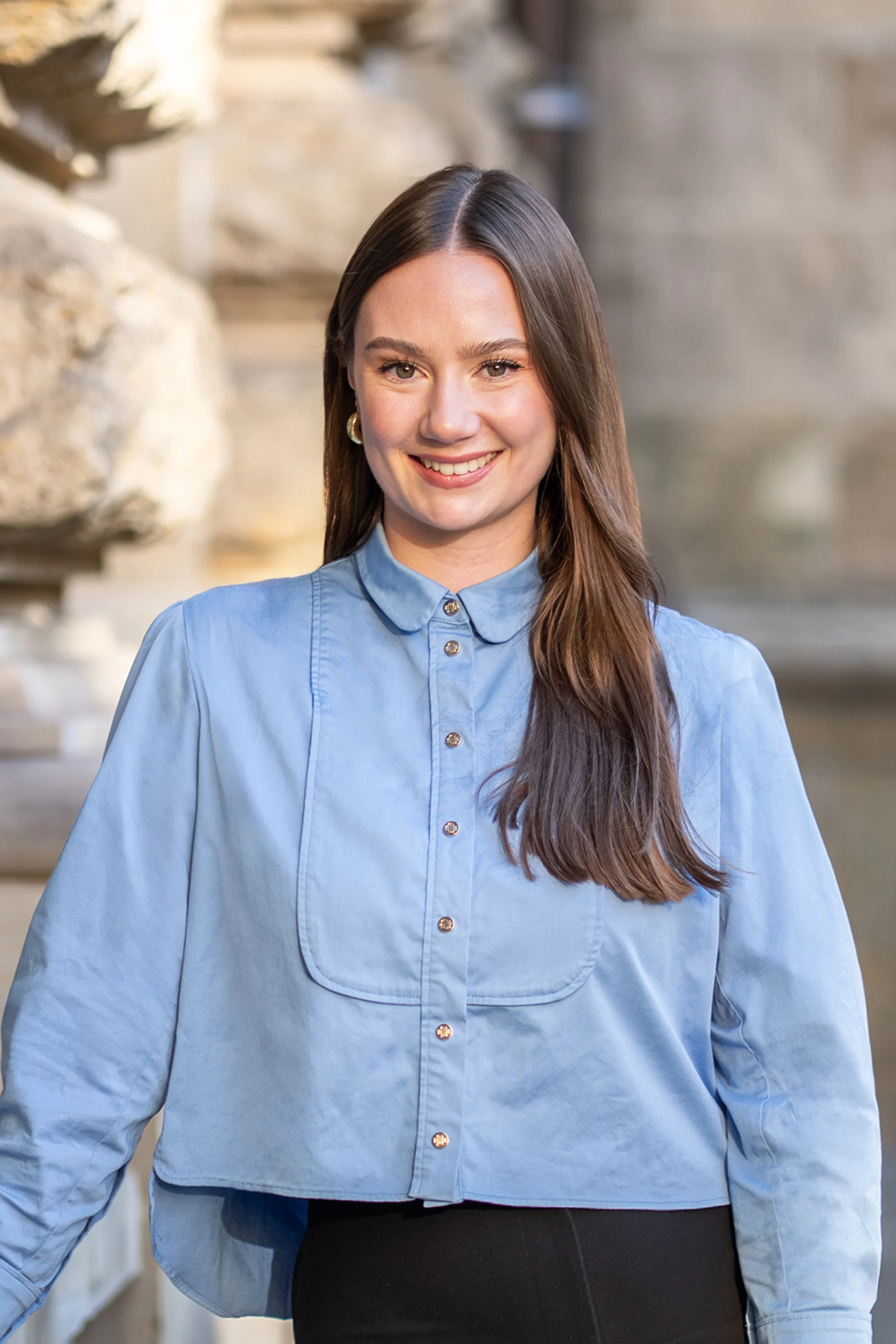 Eine Frau mit langem braunem Haar trägt eine hellblaue Bluse und steht draußen.