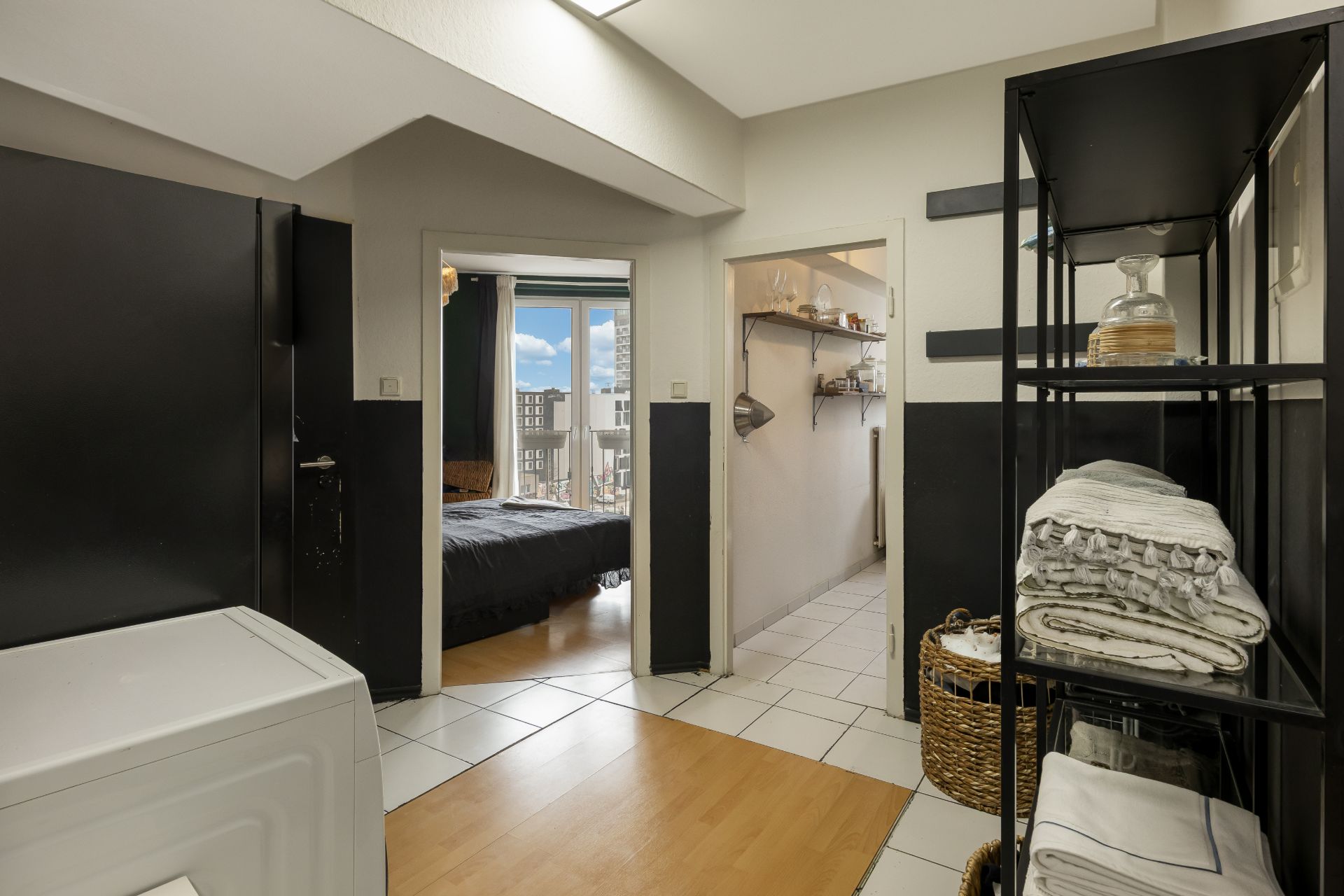 Flur mit Regal, Wäschekorb und Zugang zu Schlafzimmer und Küche in einer Wohnung.