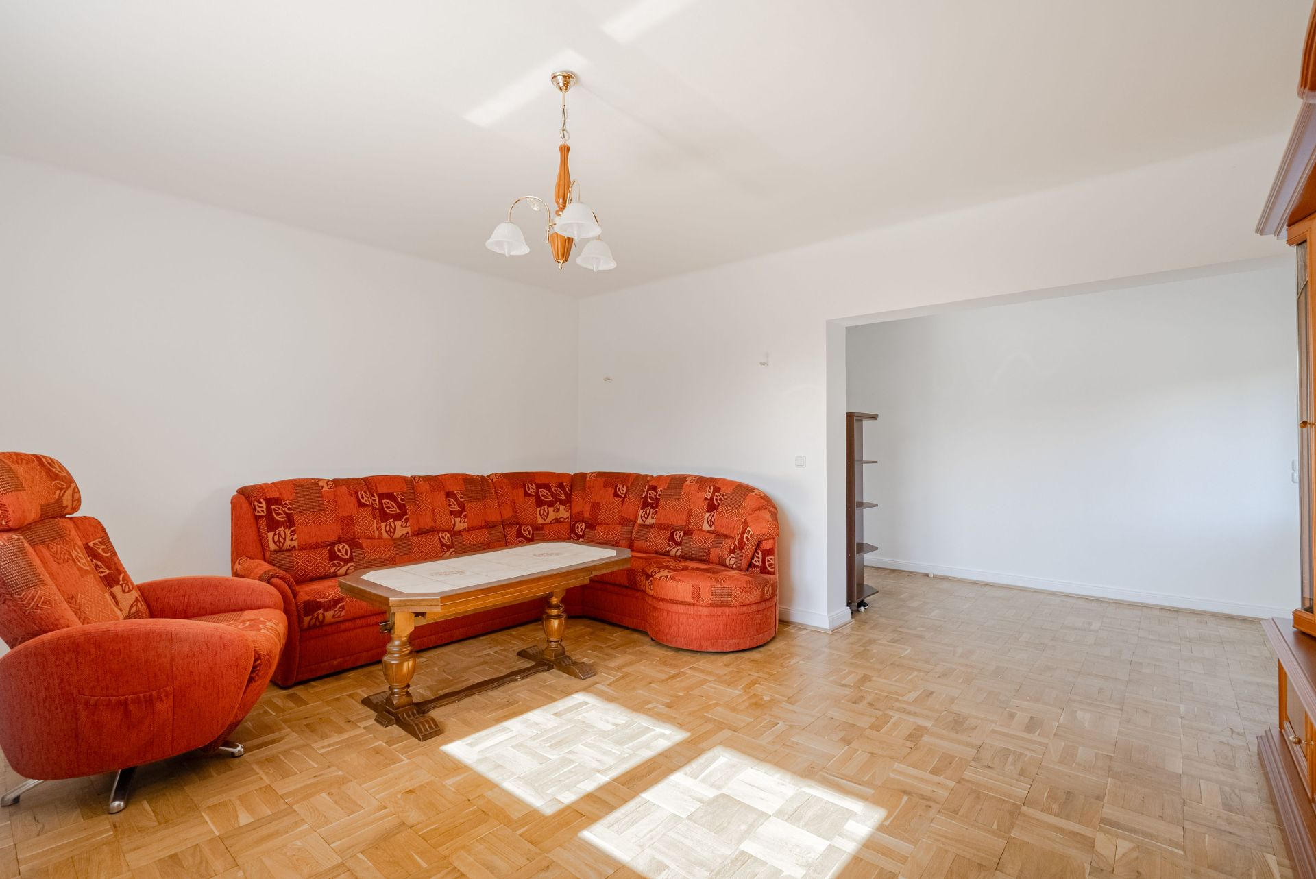 Wohnzimmer mit rotem Sofa, Sessel, Holztisch und Parkettboden, nebenan ein leerer Raum.