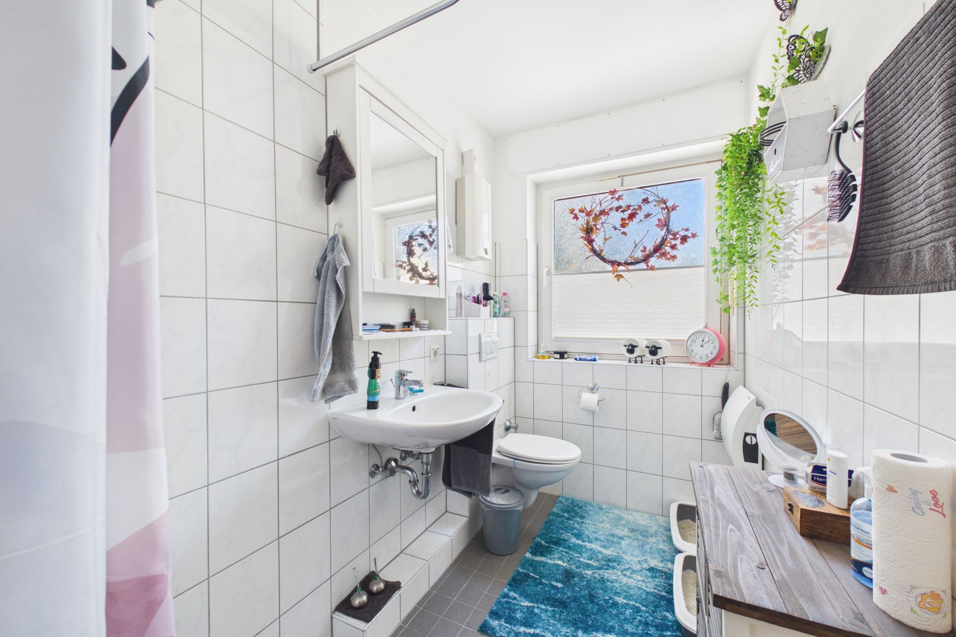 Helles Badezimmer mit Fenster, Waschtisch, Spiegel, Toilette und Regalen, türkisfarbener Teppich am Boden.