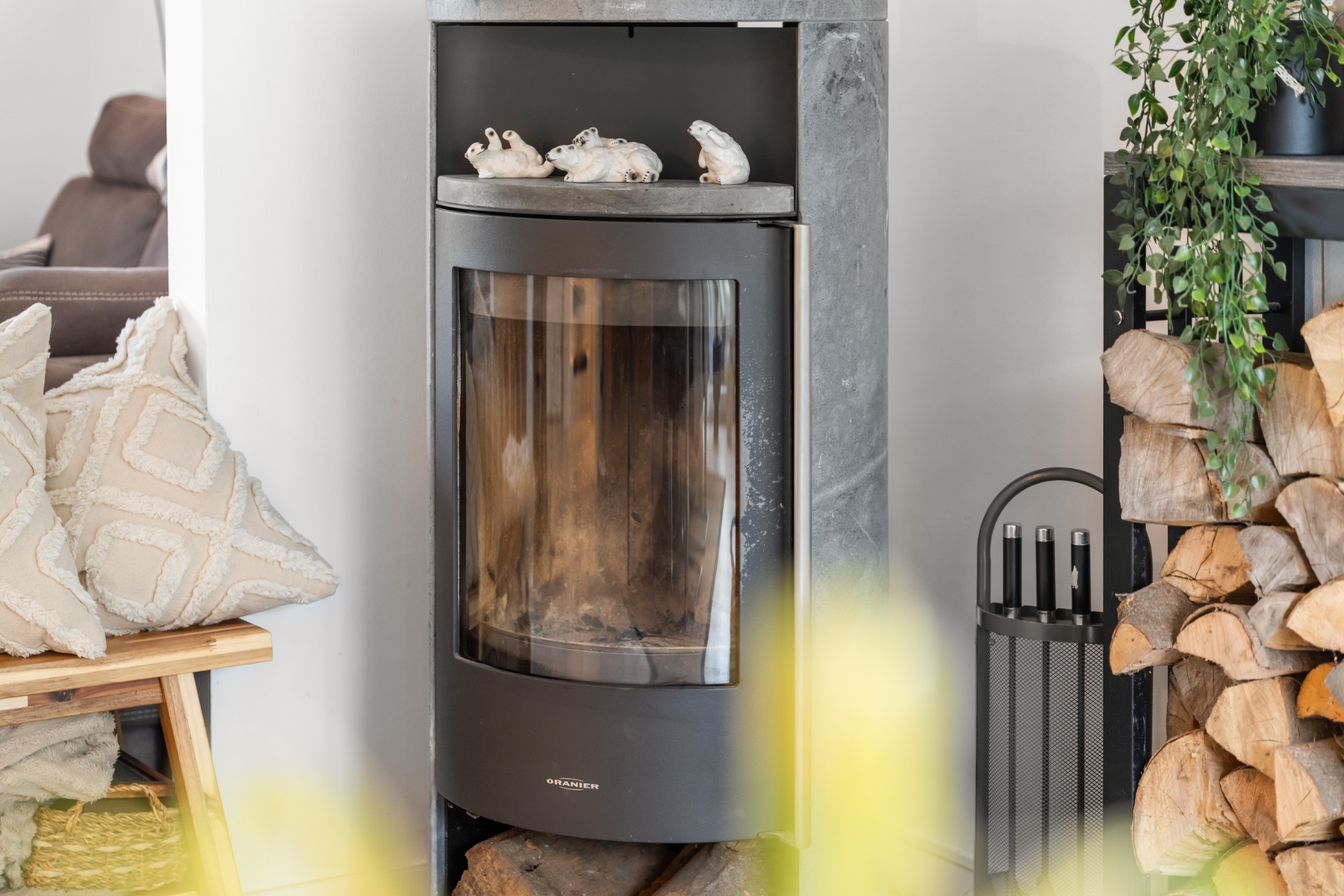 Moderner Kaminofen mit Dekofröschen, daneben ein Holzregal und Kissen auf einer Bank.