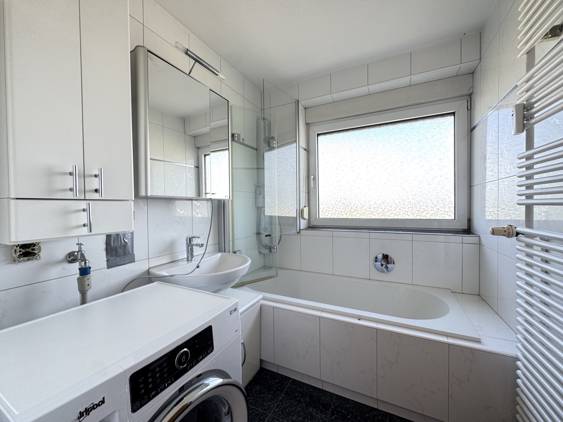 Helles Badezimmer mit Badewanne, Waschmaschine, Waschbecken, Spiegel und Fenster.