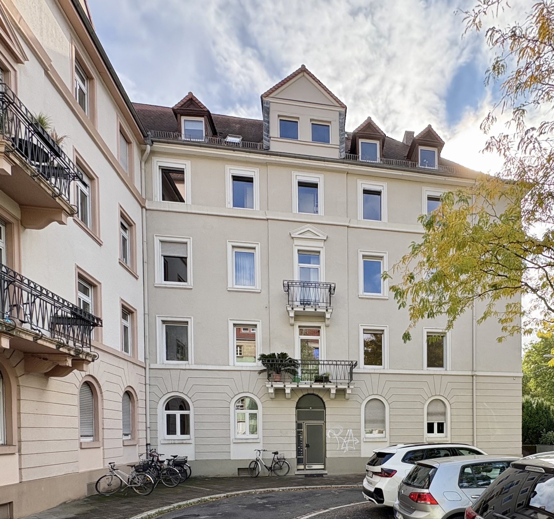 Mehrstöckiges Wohnhaus mit Balkonen, geparkten Autos und Fahrrädern vor dem Eingang.