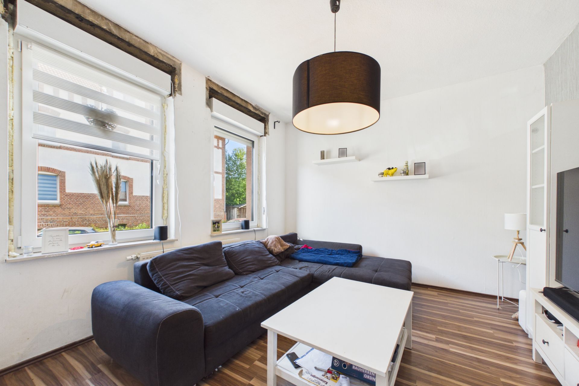 Wohnzimmer mit dunklem Ecksofa, weißem Tisch, Holzfußboden und großen Fenstern.