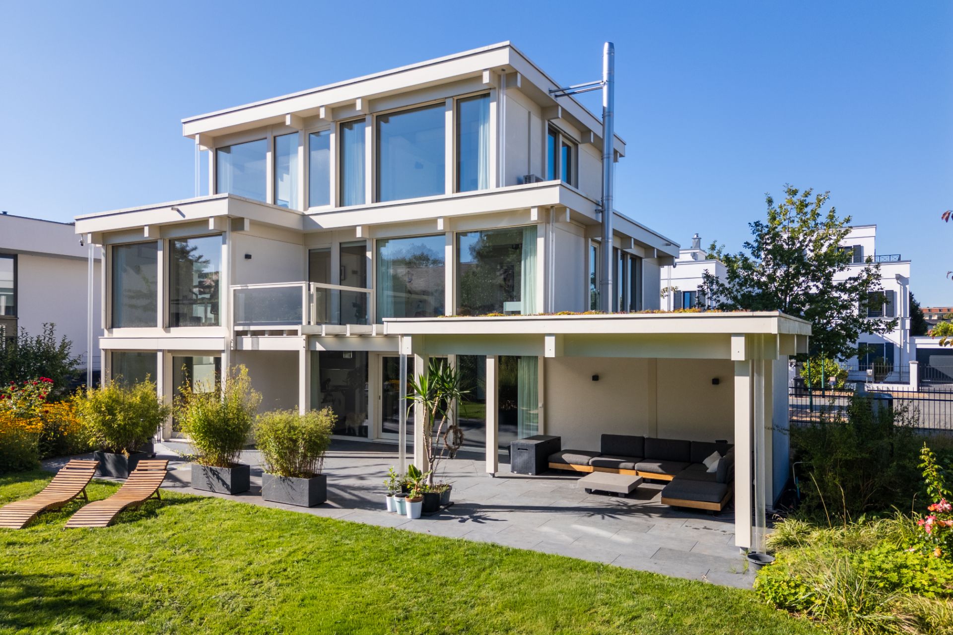 Modernes Wohnhaus mit großer Fensterfront, Terrasse, Gartenmöbeln und gepflegtem Rasen im Vorgarten.
