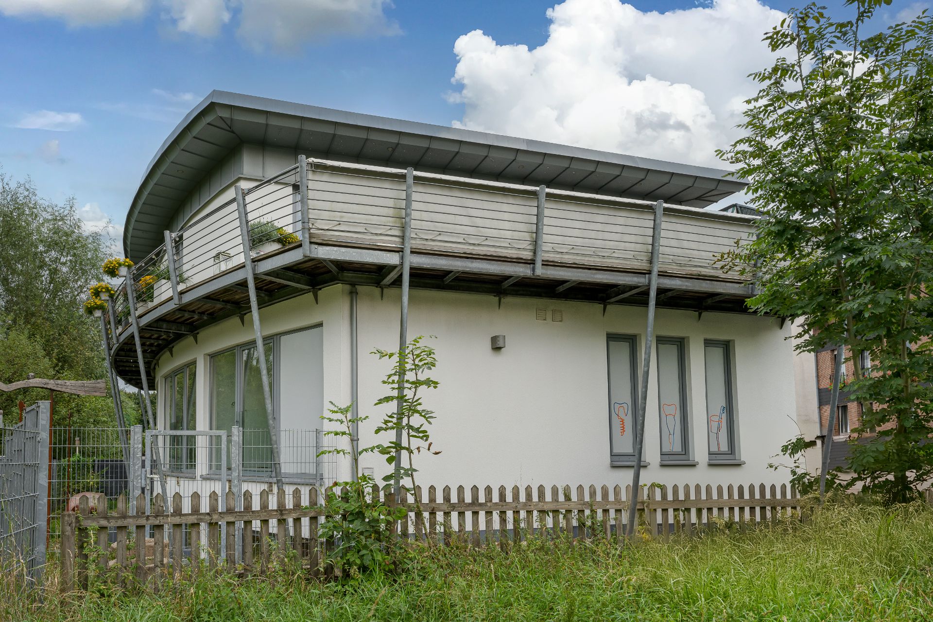 Modernes, rundes Haus mit Balkon, Metallstützen und kleinem Holzzaun im Vorgarten.
