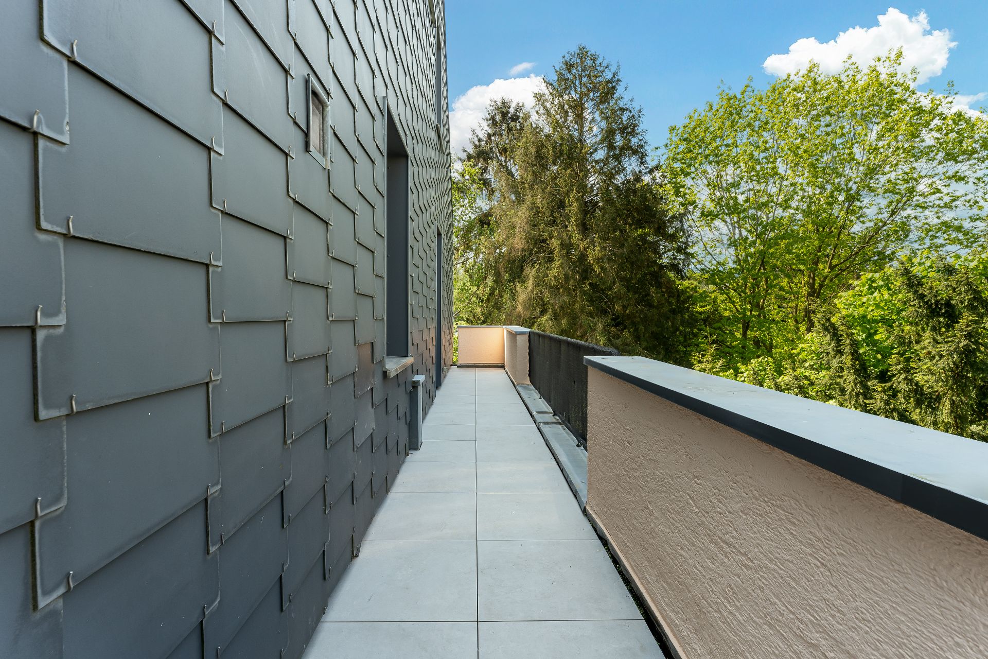 Langer Balkon mit grauen Fliesen, links Hauswand, rechts Blick auf Bäume.