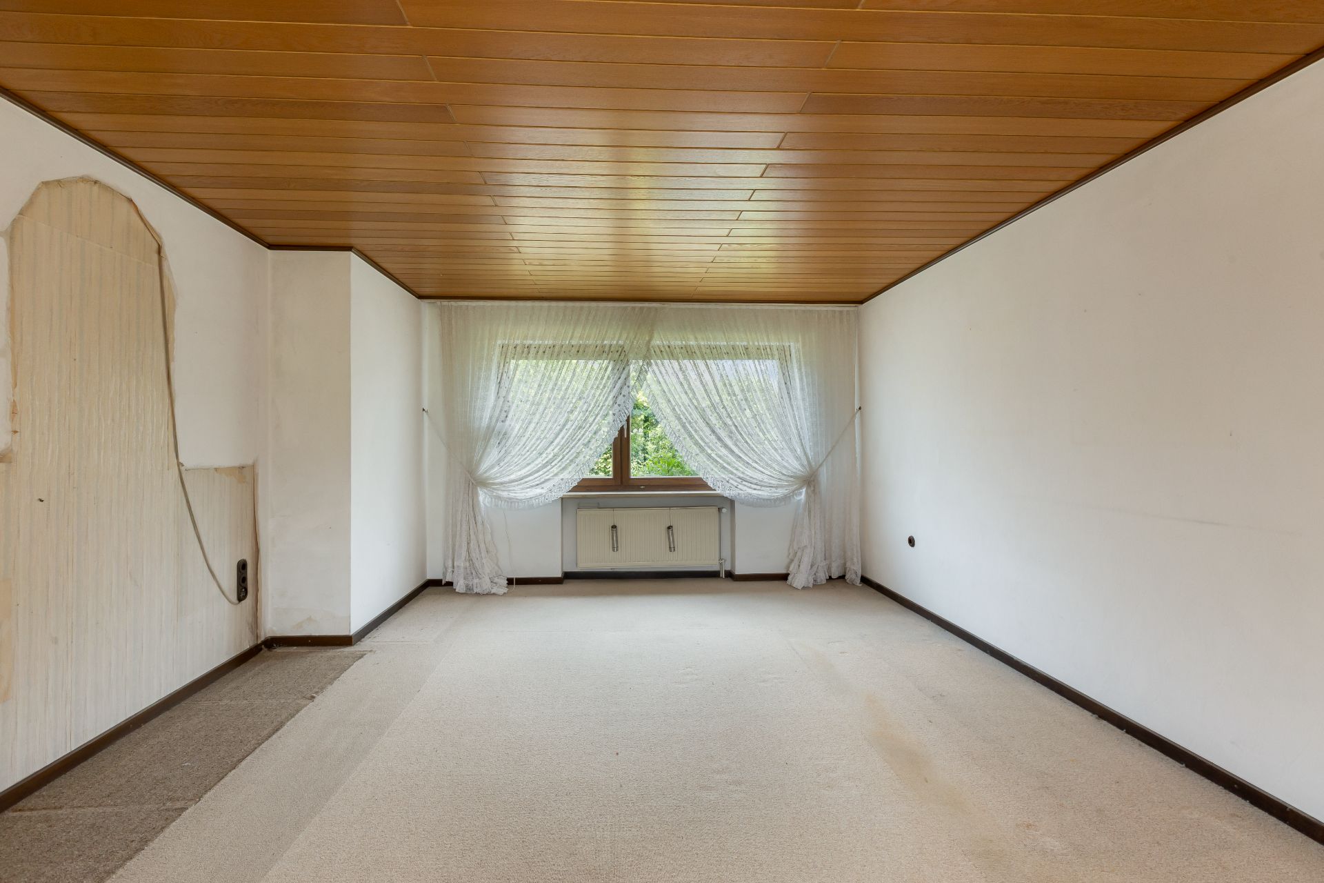 Leerer Raum mit Holzdecke, Teppichboden, großem Fenster und weißen Vorhängen.