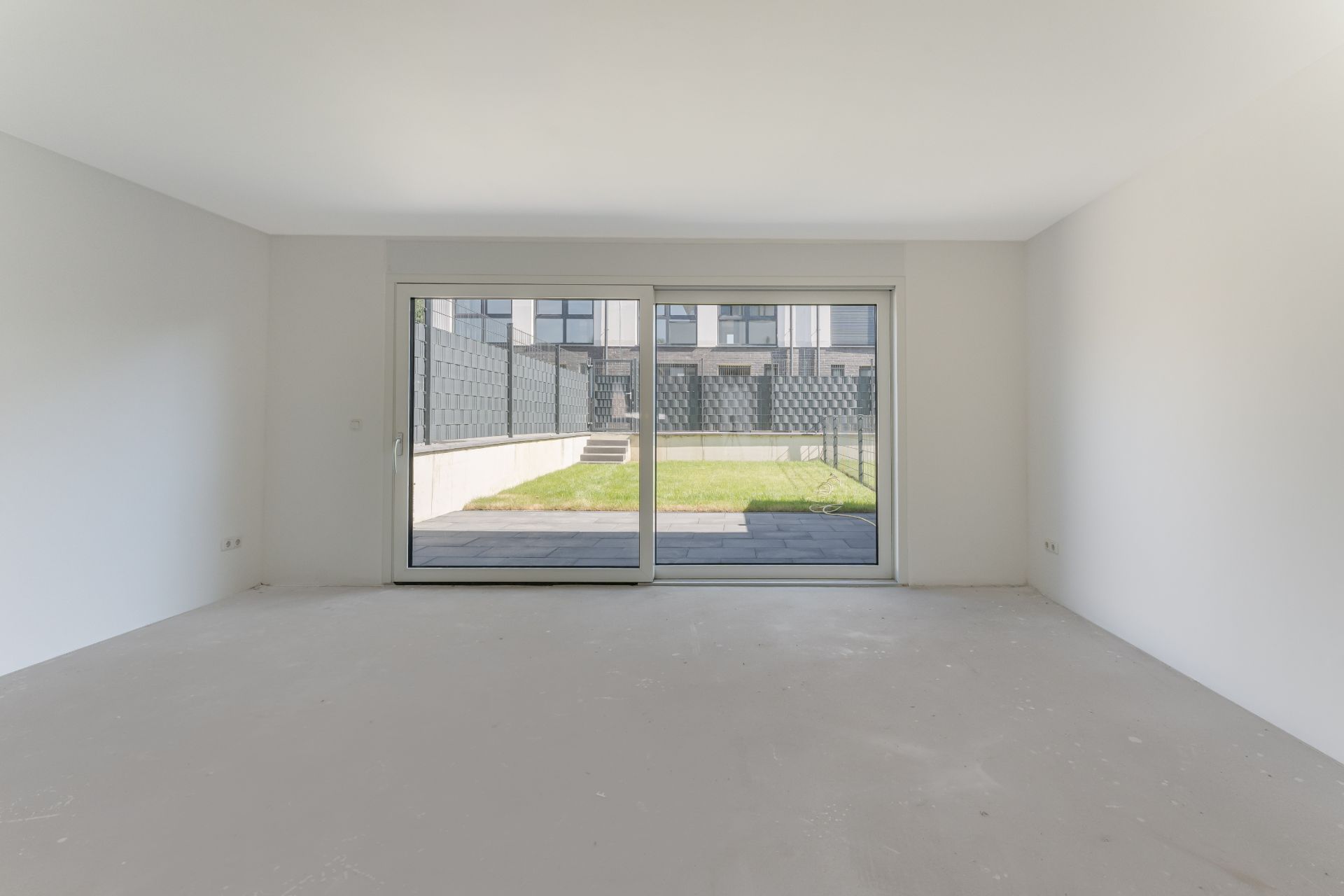 Leeres Zimmer mit großen Glas-Schiebetüren und Blick auf einen Garten.