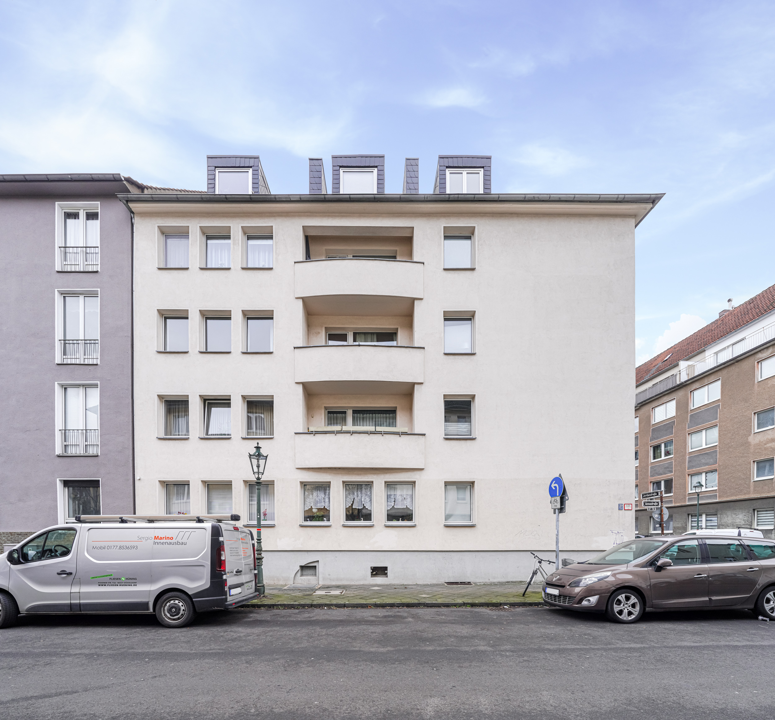 Vorderansicht eines mehrstöckigen Wohnhauses mit Balkonen und geparkten Autos auf der Straße.
