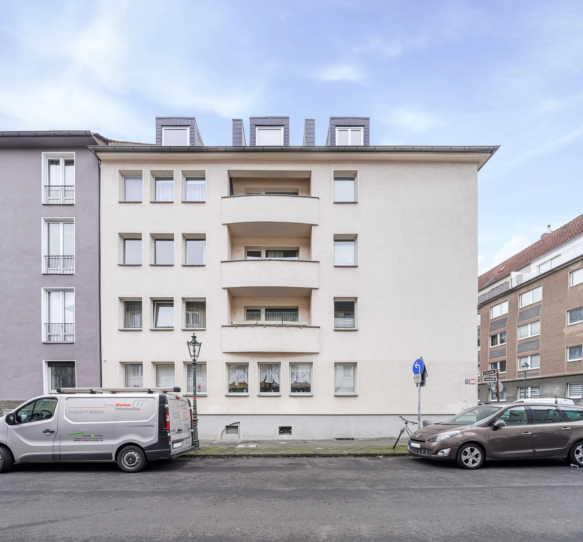 Vorderansicht eines mehrstöckigen Wohnhauses mit Balkonen und geparkten Autos auf der Straße.