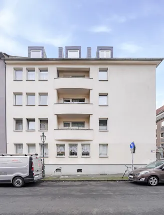 Vorderansicht eines mehrstöckigen Wohnhauses mit Balkonen und geparkten Autos auf der Straße.