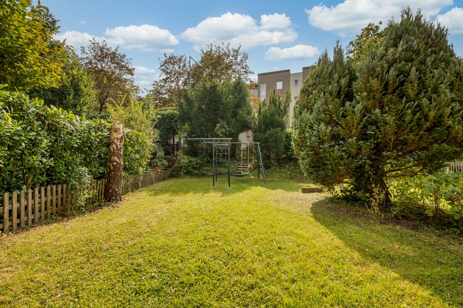 Grüner Garten mit Schaukelgestell, Rasenfläche und Bäumen, umgeben von Holzzaun.