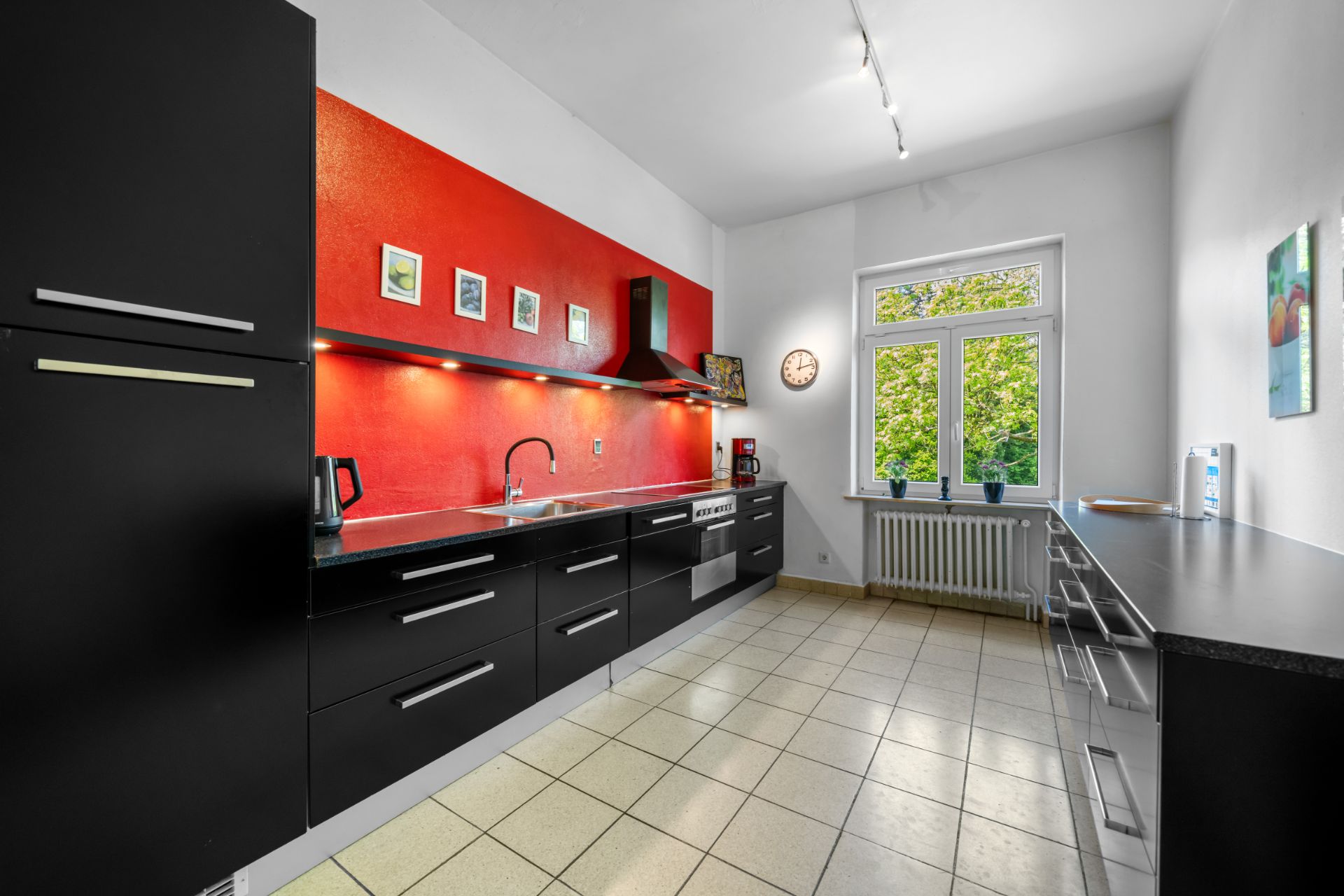 Moderne Küche mit schwarzen Schränken, roter Wand und großem Fenster mit Ausblick auf grüne Bäume.