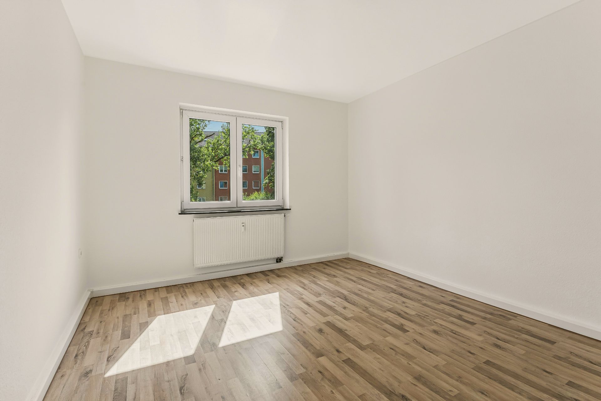 Leeres Zimmer mit Holzfußboden, weiß gestrichenen Wänden und Fenster mit Blick nach draußen.