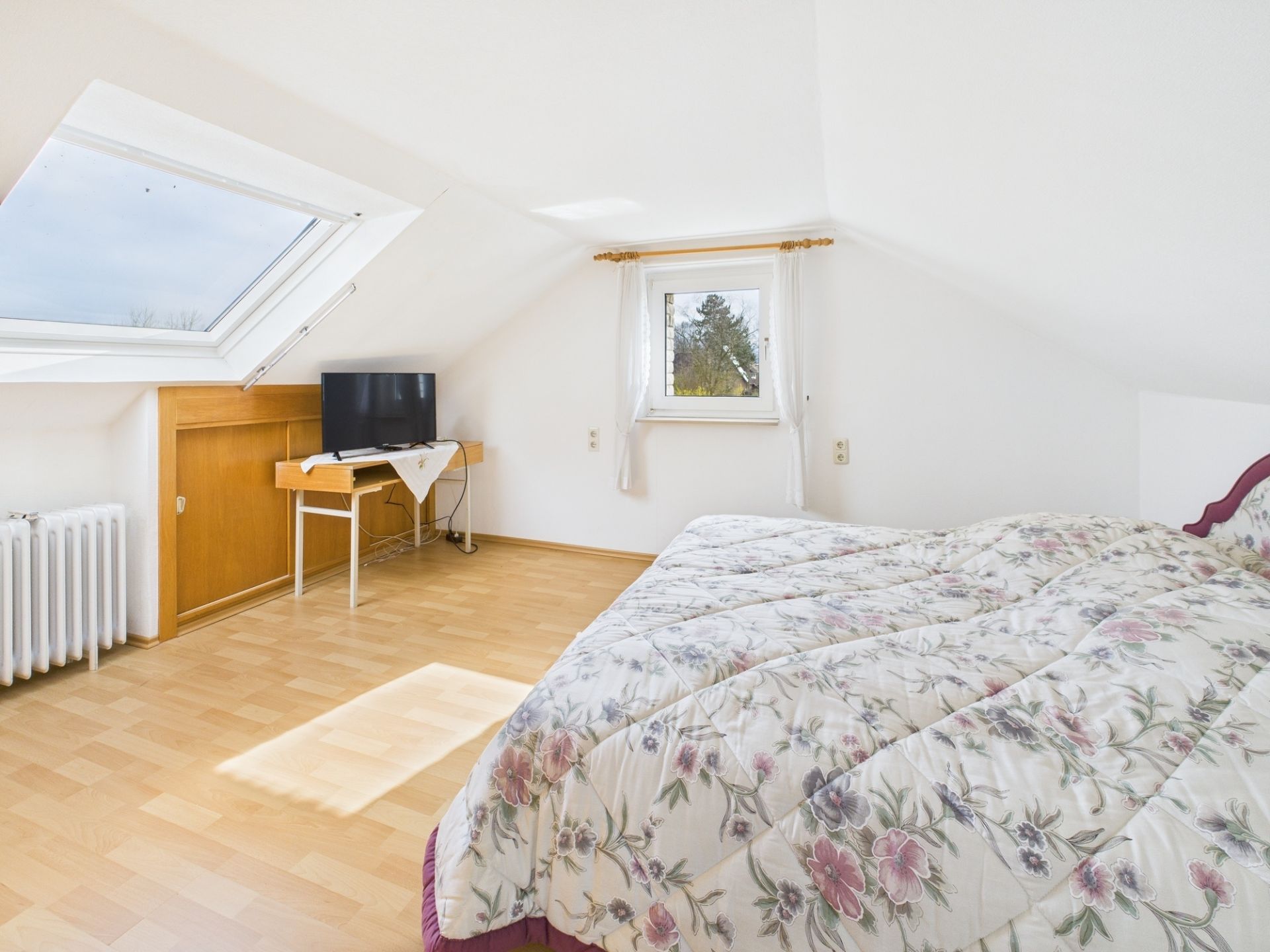Helles Schlafzimmer mit Dachfenster, Bett mit Blumenbettdecke und Fernseher auf einem kleinen Tisch.