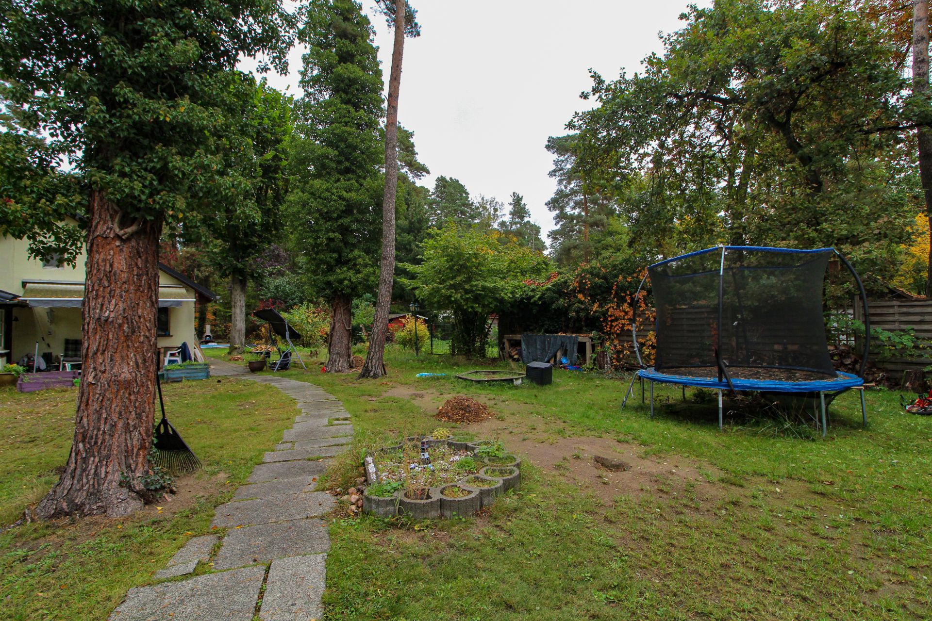 Garten mit Bäumen, Trampolin, Rasen, Steinweg und Haus im Hintergrund.