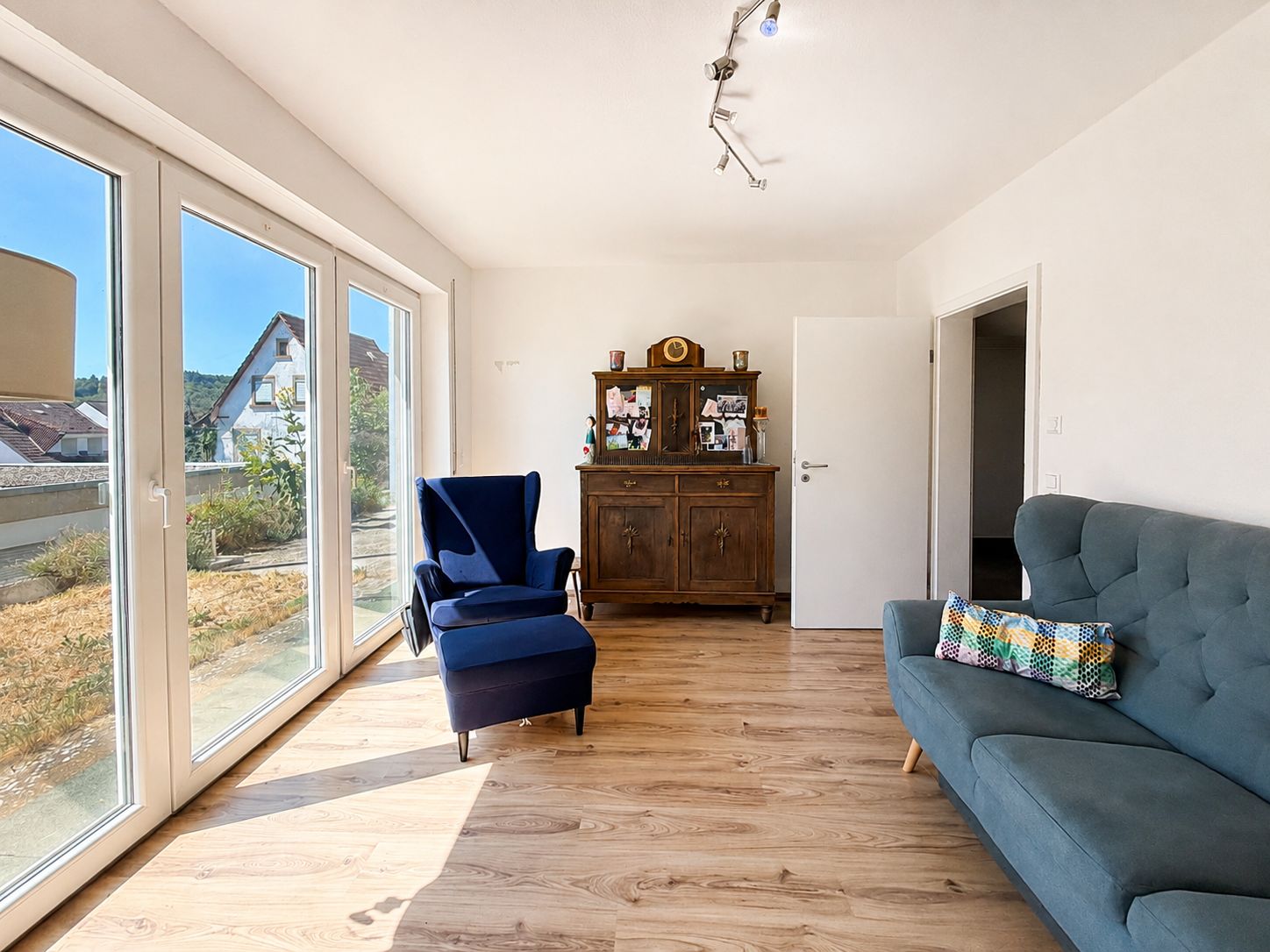 Helles Wohnzimmer mit großem Fenster, blauem Sessel, grauem Sofa und Holzschrank auf Parkettboden.