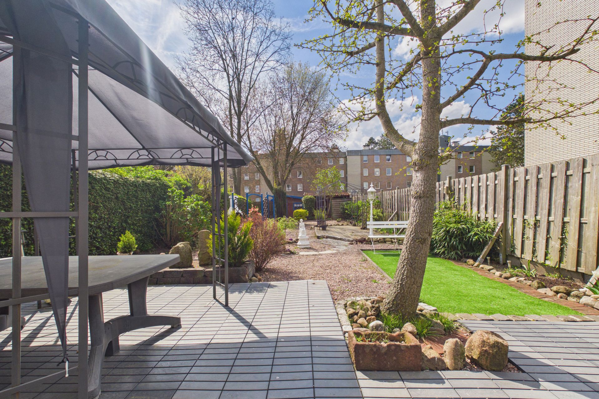 Gepflasterter Garten mit Pavillon, Tisch, Baum und Zaun, dahinter Wohnhäuser.