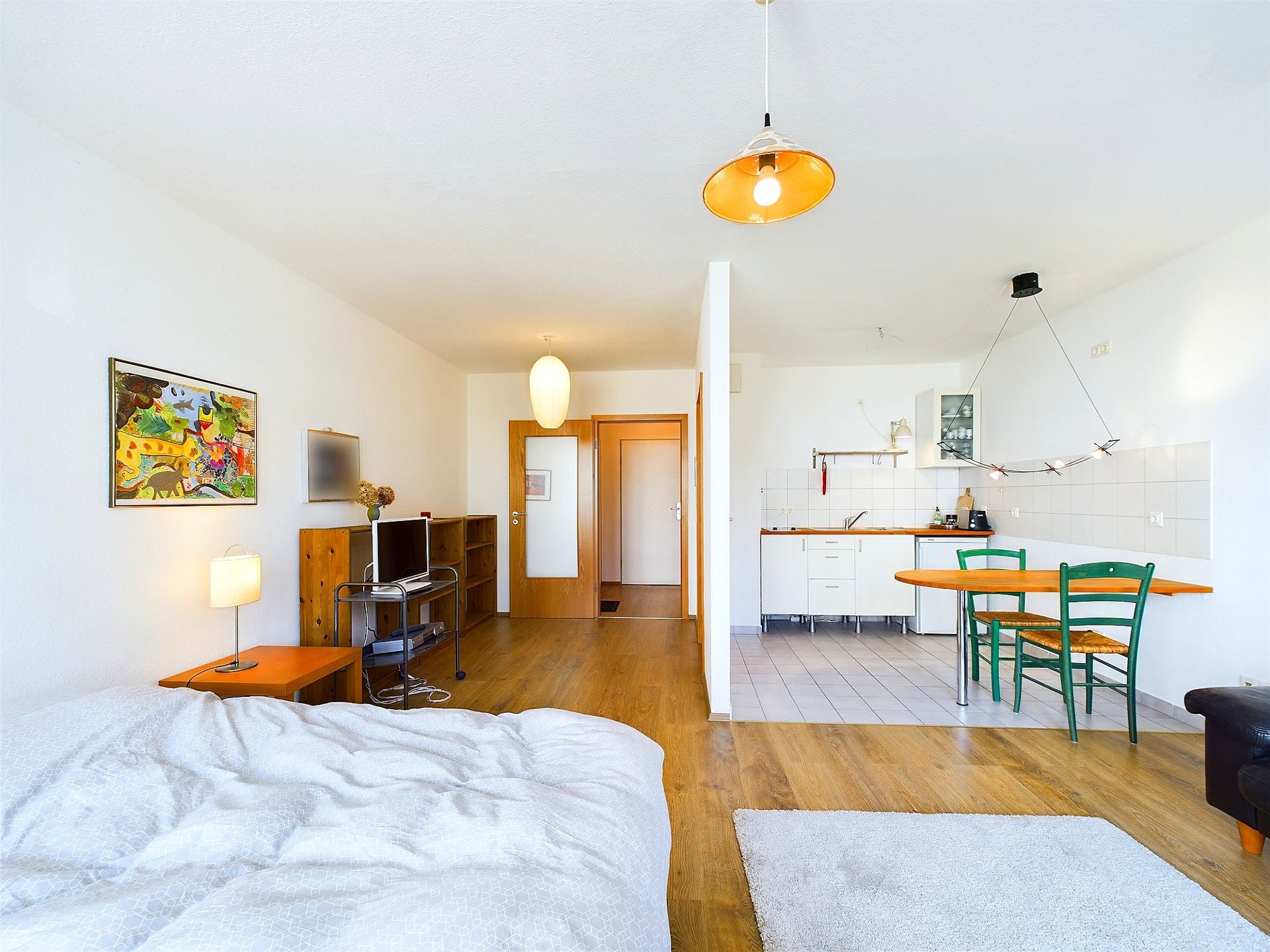 Helles Wohn- und Schlafzimmer mit Bett, Küchenecke und Esstisch mit grünen Stühlen.