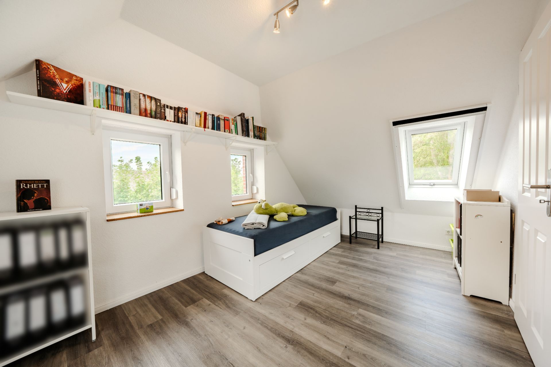 Helles Zimmer mit Einzelbett, Bücherregal, zwei Fenstern und Laminatboden.