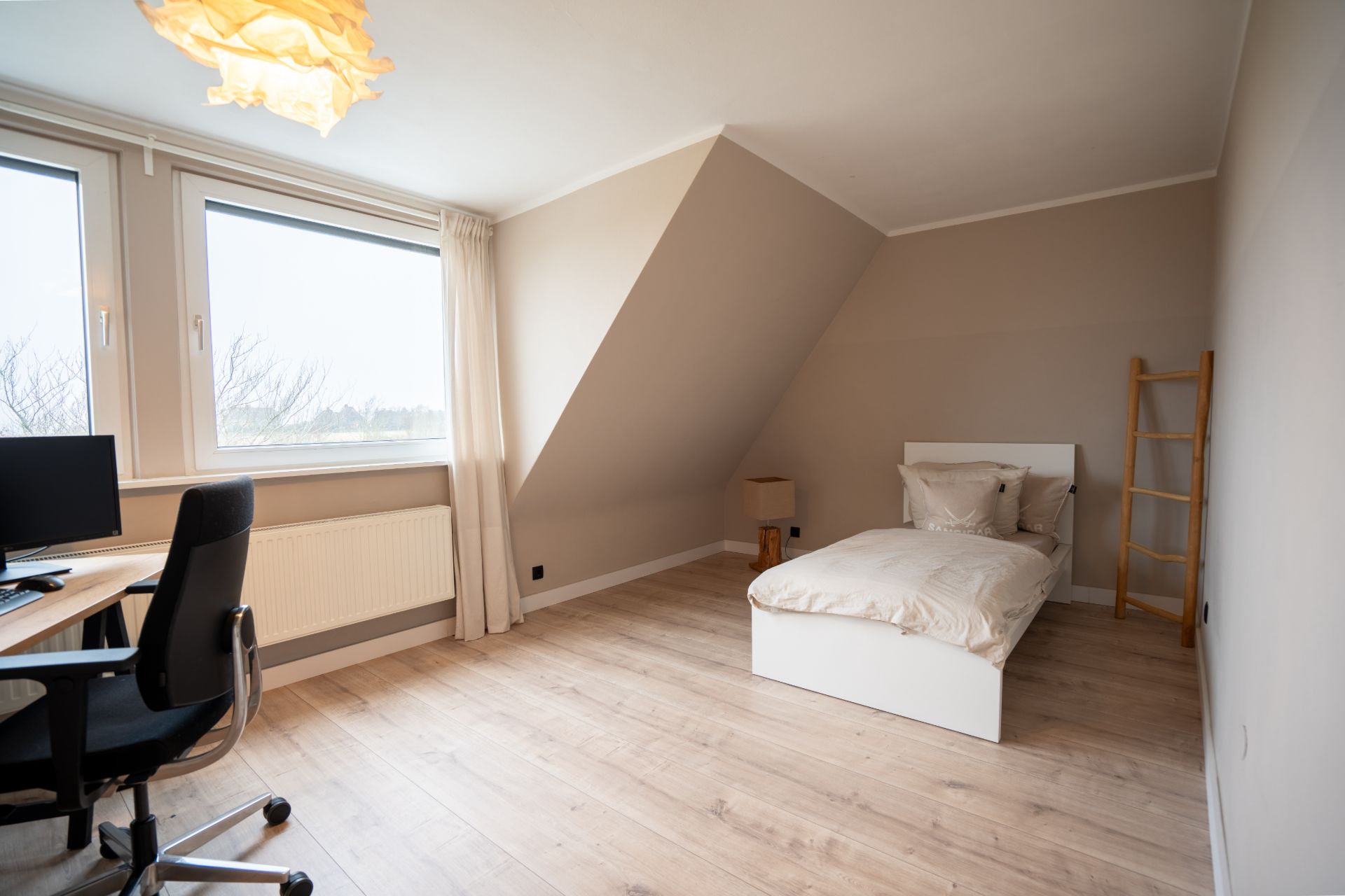 Helles Schlafzimmer mit Einzelbett, Schreibtisch, Stuhl und zwei großen Fenstern.