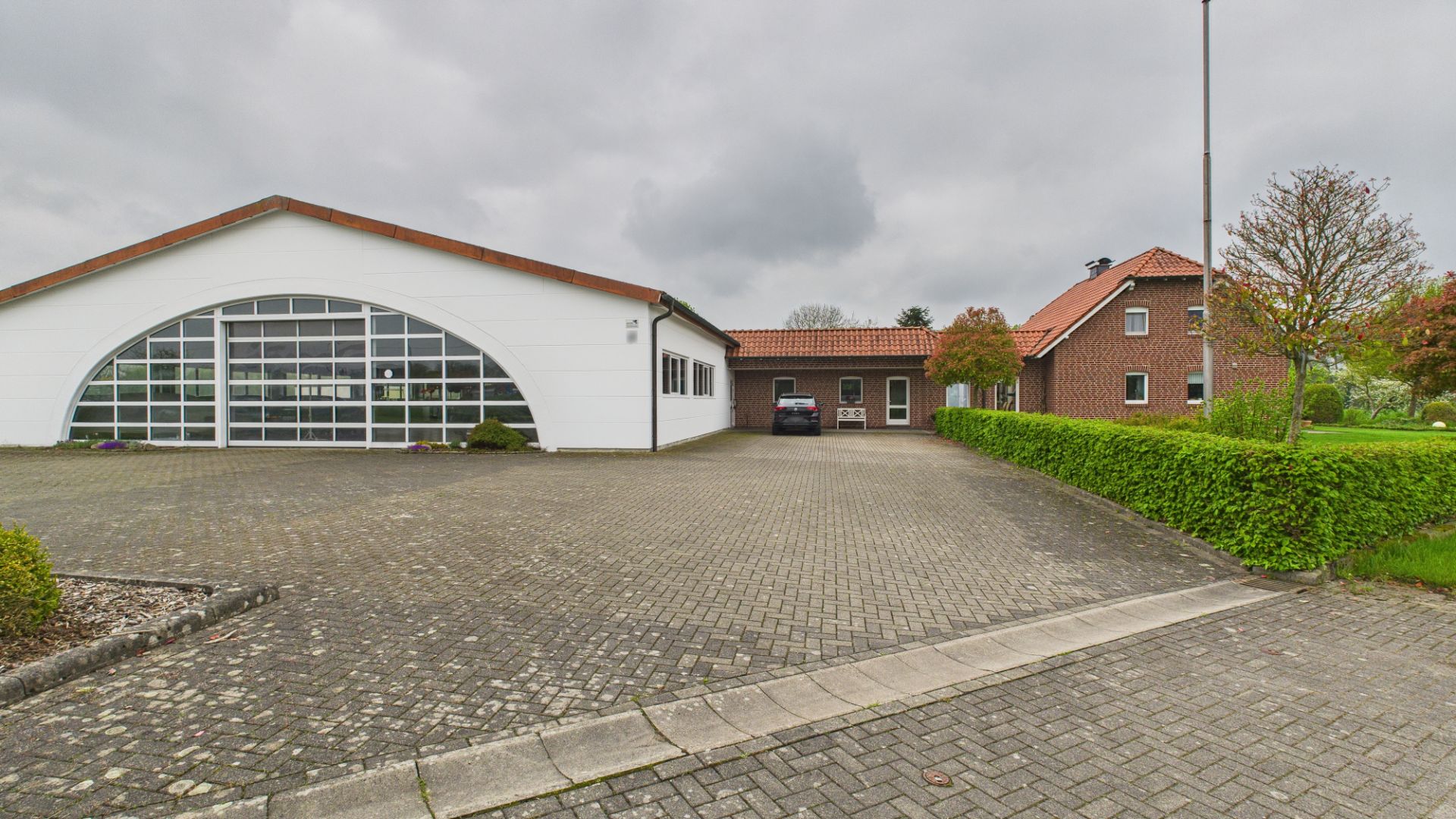 Gepflasterter Hof vor zwei Wohngebäuden mit Bäumen und Hecken bei bewölktem Himmel.