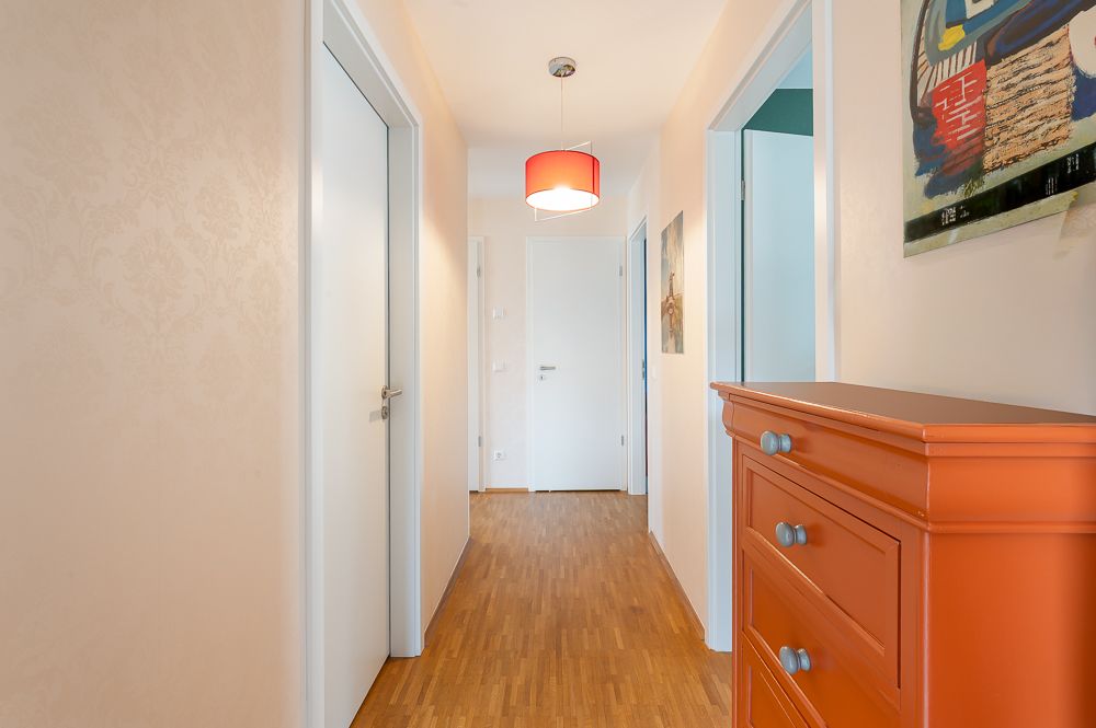 Heller Flur mit Holzfußboden, oranger Kommode, weißen Türen und roter Deckenlampe.