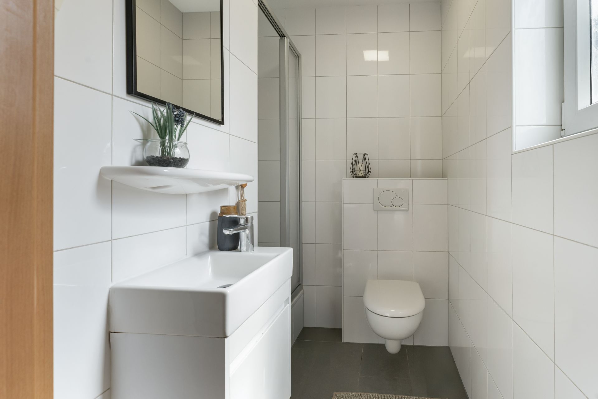 Modernes, weiß gefliestes Badezimmer mit Waschbecken, Spiegel, WC und Fenster.