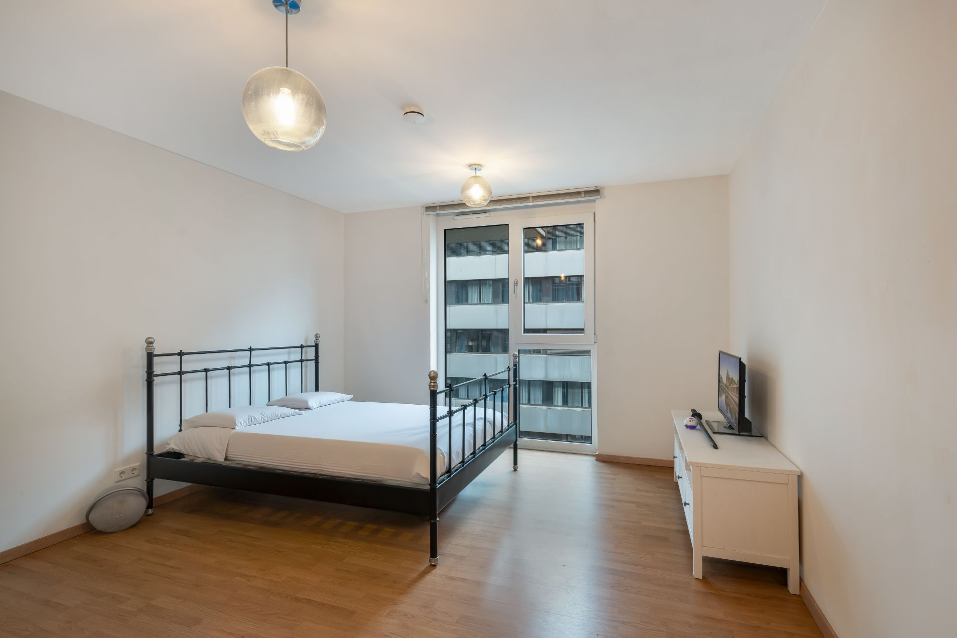 Helles Schlafzimmer mit Metallbett, Holzfußboden, Kommode und Fenster zur Straße.