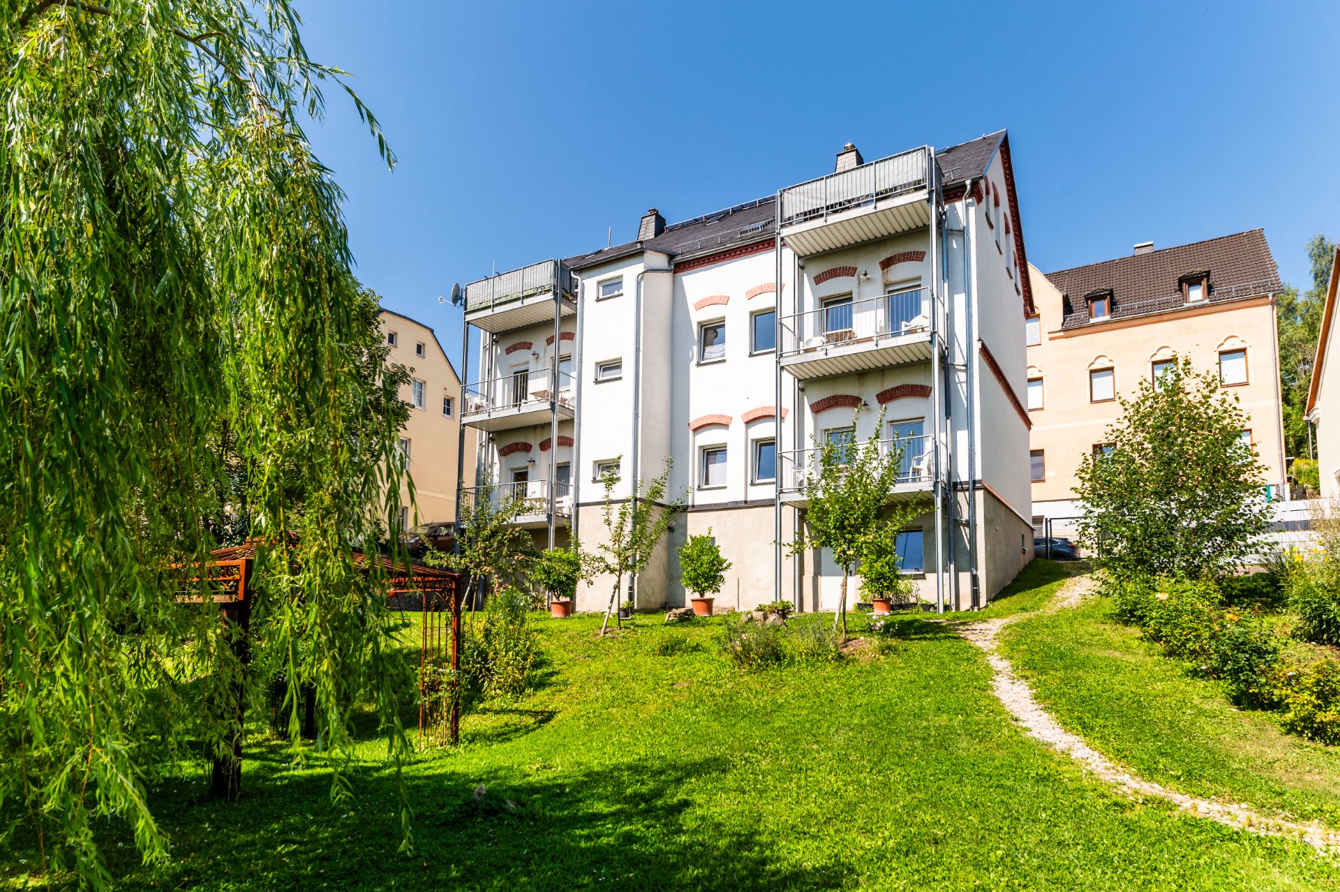 Mehrfamilienhaus mit Balkonen, großer grüner Garten und Bäumen im Vordergrund bei sonnigem Wetter.